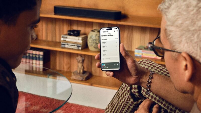 Two people with extremely 70s vibes looking at Sonos' app, with shag carpeting, wood paneling, and houndstooth pants in the frame.