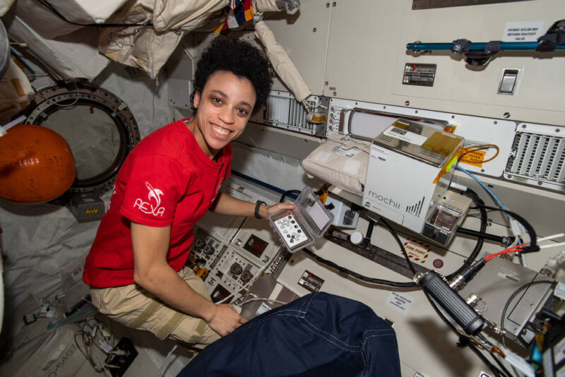 I woman holds a handheld device in front of a rack of equipment.