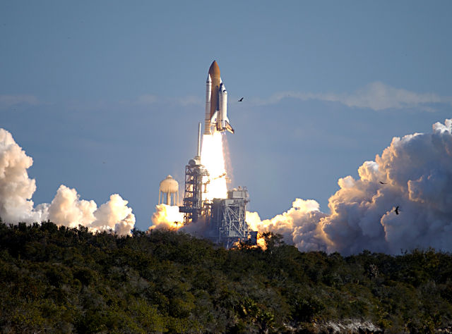 Through a cloud-washed blue sky above Launch Pad 39A, Space Shuttle <em>Columbia</em> hurtles toward space on mission STS-107. “><figcaption class=