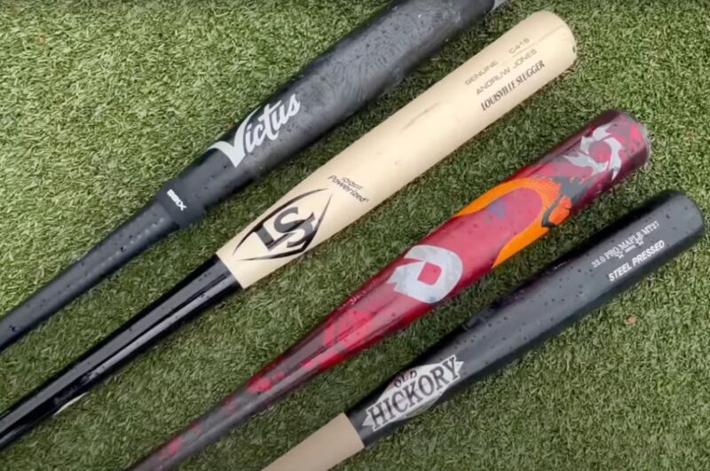 four different kinds of wood and metal bats laid flat on baseball diamond grass