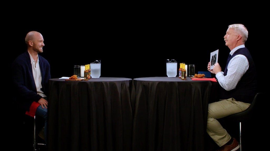 Two men sitting at a table with hot sauce and chicken wings