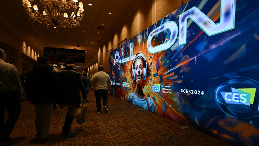 CES 2024 attendees walking by a big digital billboard that reads "ALL ON" over a photo of woman with headphones. Fire explosion behind her head.