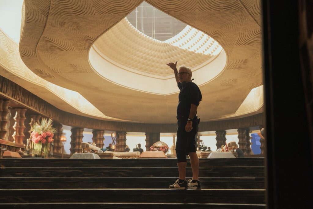 Man standing on steps of a set with a very high ceiling and teardrop shape cut out of the ceiling