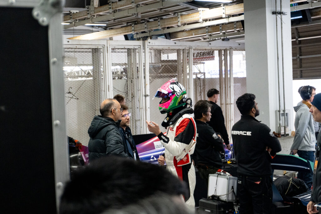 A racing driver and some engineers stand around in a garage at a track