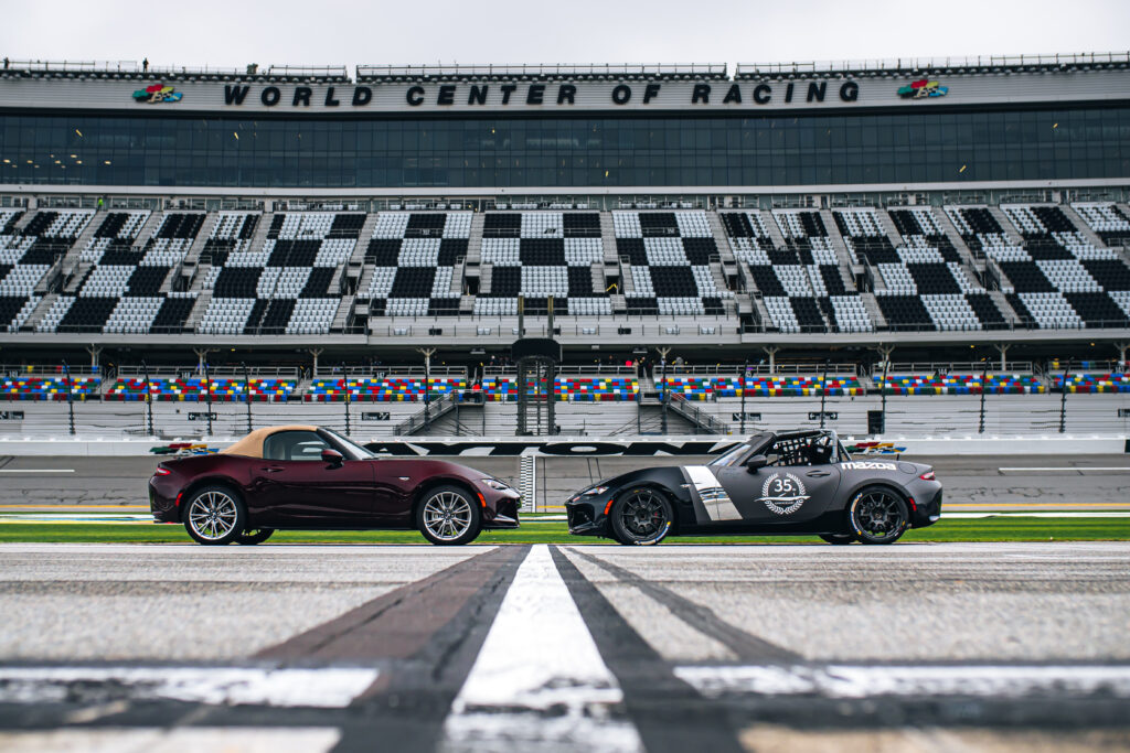 A red Mazda MX-5 street car faces a Mazda MX-5 race car, parked in front of the grandstand at Daytona