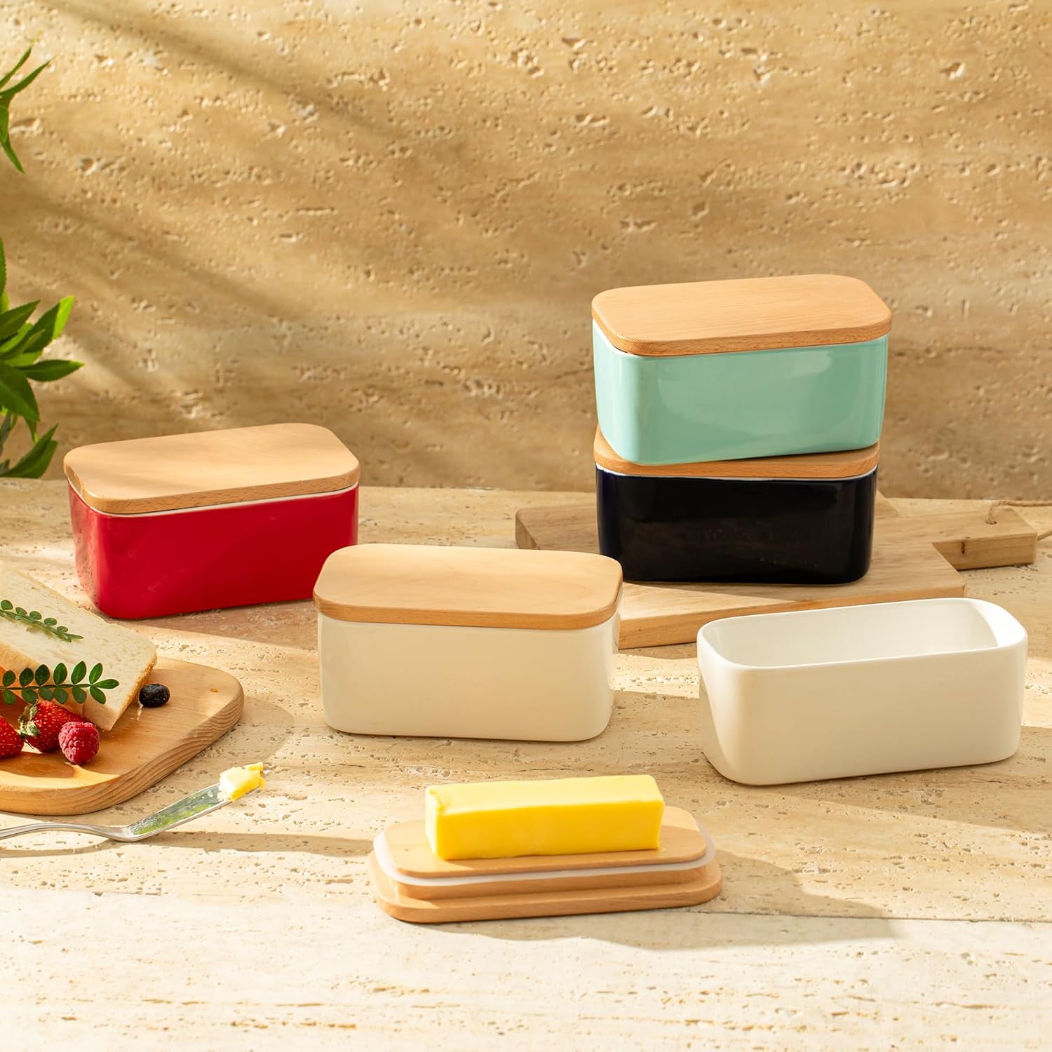 Several butter dishes with differently colored sides and a wooden top, with a stick of butter in front.