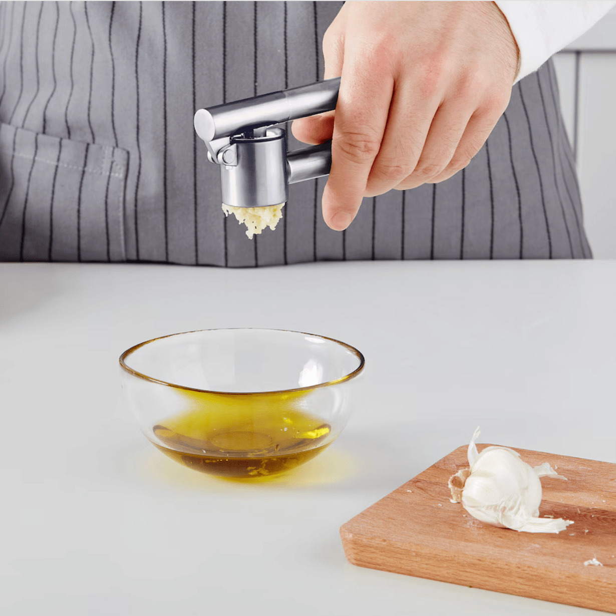 Hand of person holding a garlic press over a small glass bowl near a wooden chopping board holding with garlic on it.