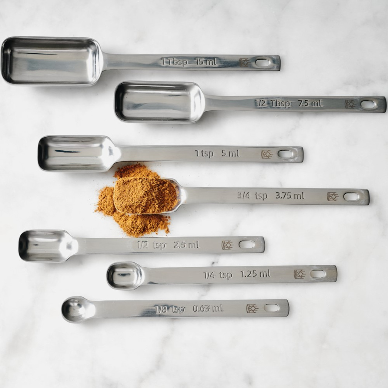 7 measuring spoons of different sizes lying parallel on a service; the center spoon is full of a brown spice.