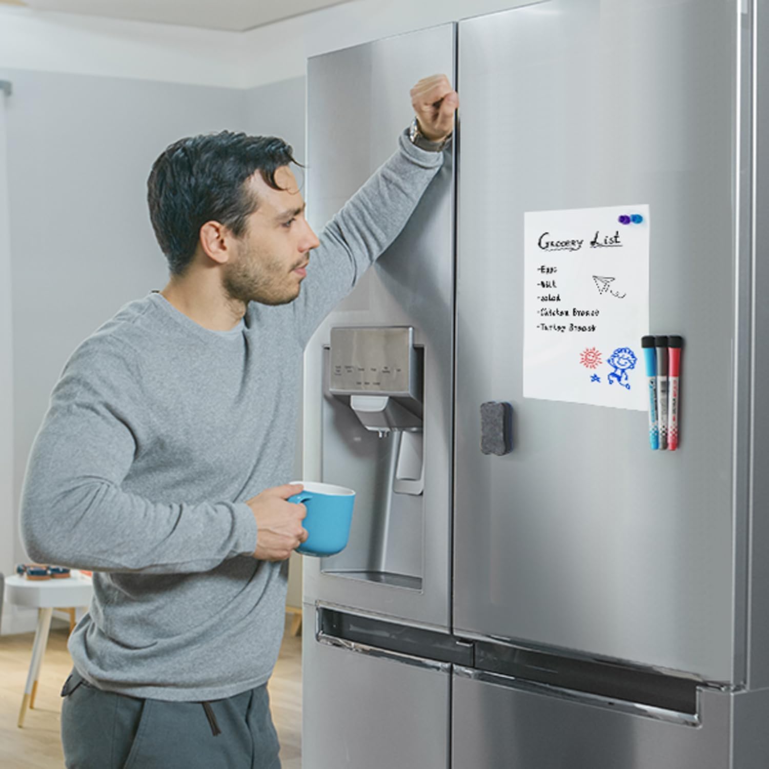 Man holding coffee leaning against refrigerator and looking at small whiteboard with list on it.